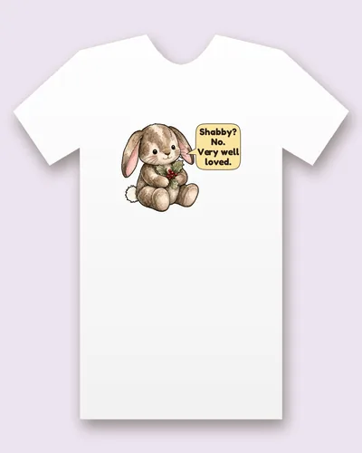 Velveteen Rabbit tee mockup: A worn, patched plush rabbit with long floppy ears, holding a sprig of holly with red berries, saying “Shabby? No. Very well loved.”